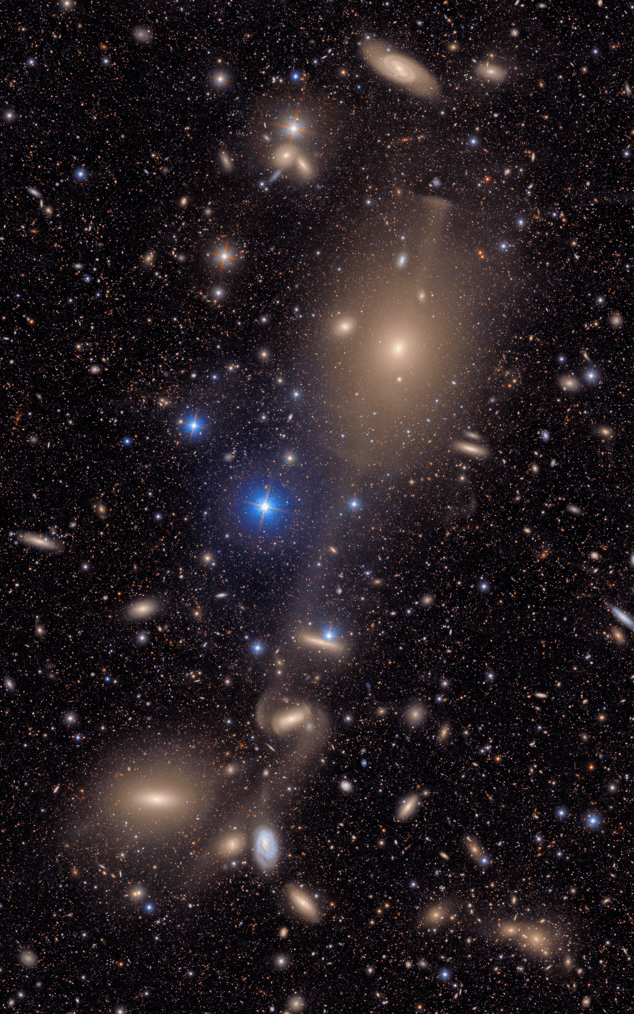 A sprawling, textured field of galaxies scattered across the deep black of space. It is filled with the delicate smudges and glowing cores of galaxies of many shapes, sizes and colors, as well as the bright multi-colored points of stars. The image focuses on a collection of interacting galaxies connected by delicate streams of stars. At top center lies a large elliptical galaxy that is dense and smooth, like a polished stone glowing with golden light. Like delicate spider silk or stretched taffy, these stellar bridges link the large elliptical to the few larger galaxies beneath, evidence of past collisions. All throughout the image, thousands of galaxies gather in clusters or are spread throughout, like glittering gems strewn on a table. Some are sharp-edged and spiral, like coiled ribbons; others round and diffuse, like polished pebbles. Still others are just smudges of various colors against the black of space. The background is peppered with pinpoint stars in reds, yellows, and blues, crisp against the velvet black.