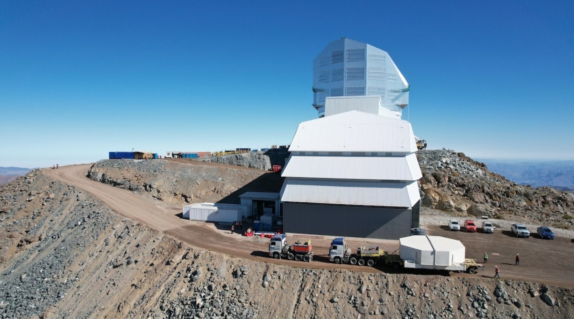 Two large hauling trucks pull a 28-foot-wide, white hexagon-shaped box on a flat trailer up a grated dirt desert road. The white hexagonal box holds Rubin Observatory’s 8.4-meter combined primary/tertiary glass mirror.
