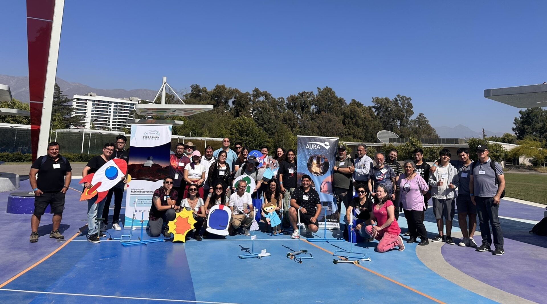 Foto grupal de treinta profesores bajo un despejado cielo azul en Chile. Algunos sostienen actividades científicas hechas a mano, como la imagen animada de un gran cohete y un casco de astronauta.